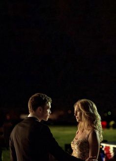 a man and woman standing next to each other in front of a field at night