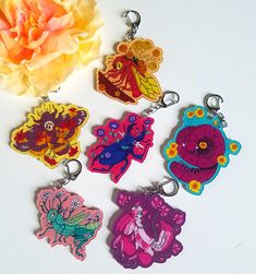 four different colored key chains sitting next to a pink flower on a white surface with an orange rose in the background