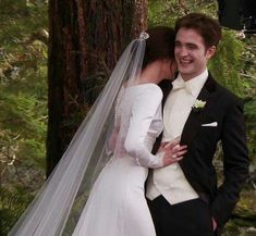 a bride and groom standing next to each other in front of a tree with their arms around each other