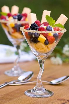 two glasses filled with fruit and cheese on top of a wooden table next to silverware