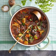 a pot filled with stew and vegetables on top of a green table cloth next to a wooden spoon