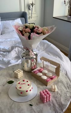 a table topped with a cake and flowers on top of a white bed covered in pillows