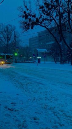 a bus driving down a snow covered road at night with buildings in the back ground