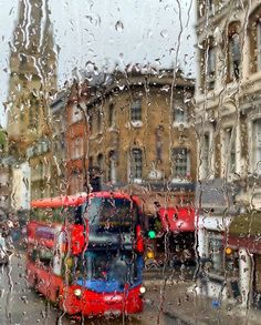 two red double decker buses passing each other on a city street through a rain soaked window