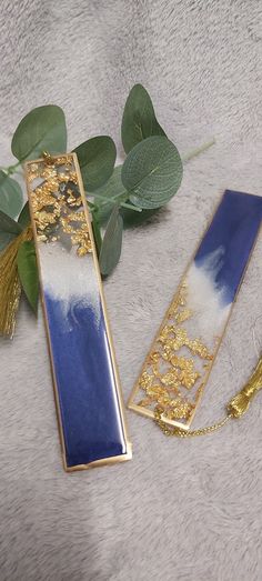 two blue and gold bookmarks sitting on top of a white blanket next to green leaves