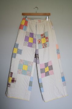 a pair of white pants with colorful patchwork on them hanging from a wooden hanger