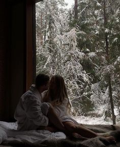 a man and woman sitting on a bed in front of a window with snow covered trees outside