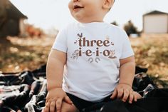 a baby sitting on top of a blanket wearing a shirt that says three ie - o