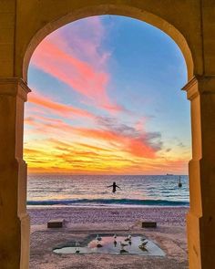 a person on a surfboard in the ocean under an arch at sunset with birds flying around