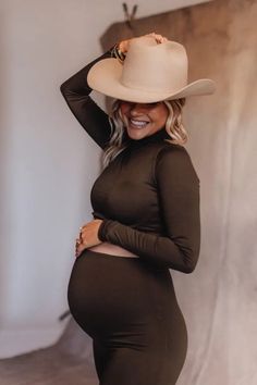 a pregnant woman wearing a brown dress and cowboy hat, posing for the camera with her hands on her head