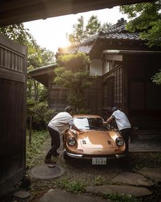 two men working on an orange sports car in front of a house with trees and bushes