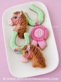 decorated cookies with horse heads and ribbons on a white platter against a pink background