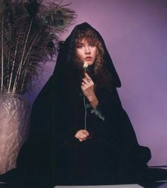 a woman in a black robe holding a flower next to a vase with feathers on it