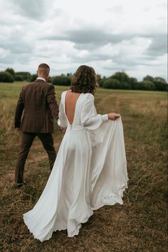 a bride and groom walking through a field together in their wedding attire, back to the camera