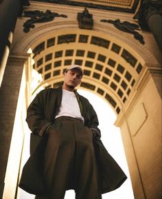 a man standing in front of an archway wearing a coat and hat with his hands on his hips