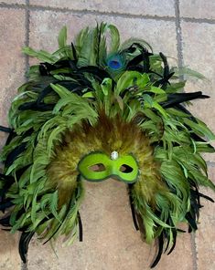 a green mask with feathers on it sitting on a tile floor next to a plant