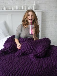 a woman sitting on a bed with a purple blanket and holding a drink in her hand