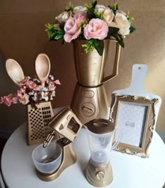 a gold blender sitting on top of a white table next to flowers and utensils