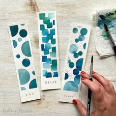 three watercolor bookmarks with blue and green designs on them, one is holding a paintbrush