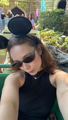 a woman wearing mickey mouse ears sitting in a green chair at an amusement park with other people behind her