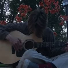 a man with long hair playing an acoustic guitar in front of some trees and flowers