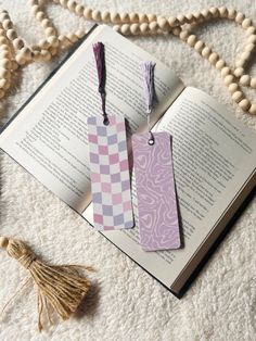 two bookmarks sitting on top of an open book next to beads and tassels