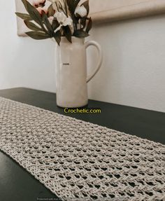 a crocheted table runner with flowers in a white vase on a black table