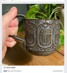 a hand holding a coffee mug on top of a wooden table next to a potted plant