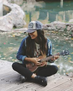 a woman sitting on the ground playing an ukulele next to a body of water