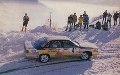 a rally car driving down a snow covered road with people standing on top of it