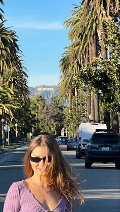 a woman wearing sunglasses standing on the side of a road with palm trees in the background