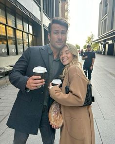 a man and woman standing next to each other on the street holding coffee cups in their hands