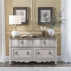 a white dresser with two pictures on the wall and a lamp in front of it