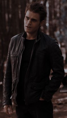 a man standing in the woods with his hand on his hip wearing a leather jacket