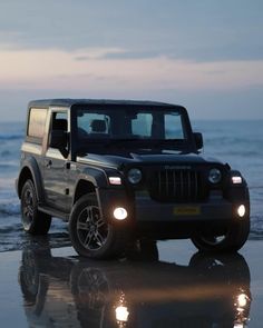 a black jeep is parked on the beach at dusk with its lights on and it's reflection in the wet sand