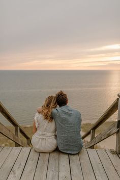 a man and woman sitting on top of a wooden deck next to the ocean at sunset