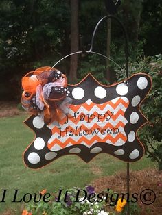 a happy halloween sign hanging from a pole in the grass with flowers and trees behind it