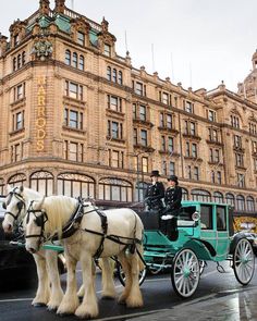 two white horses pulling a green carriage in front of a large brown building on a city street