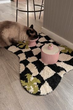 a siamese cat eating out of a bowl on the floor next to a rug