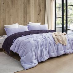 a bed with purple comforter and pillows in a room next to a window,