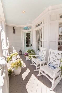 three white rocking chairs on a porch with flowers and potted plants in the foreground