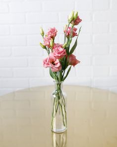 pink flowers are in a clear vase on a round table with white brick wall behind it