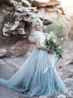 a woman in a blue and white wedding dress holding a bridal bouquet standing next to a rock formation