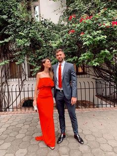 a man and woman standing next to each other in front of some trees with red flowers