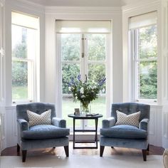 two chairs and a table in front of three large windows with flowers on the table