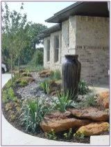 a large vase sitting in the middle of a flower bed next to a building with cars parked nearby