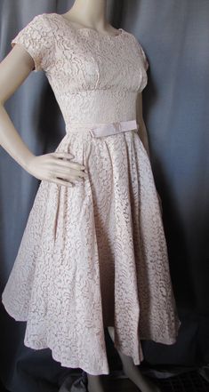 "Pretty lace party dress in a buttercream tone of beige.  Typical of late'50s, early '60s design, it has a tightly fitted empire bodice and full skirt. The waist is set slightly lower than the natural waist and is highlighted with a self fabric belt with 6\" bow at front. It has 4 1/2\" cap sleeves. The back plunges 5 1/2\" to 15\" metal zipper. The under bust seam is 9 1/2\" from throat. The dress is lined. No maker label Size: no size label--35\" bust; 27\" at the natural waist; 34\" at slight Fitted Beige A-line Lace Dress, Beige Fitted A-line Lace Dress, 1950s Style Cream Evening Dress, 1950s Cream Party Dress, 1950s Style Cream Party Dress, Cream Fitted Lace Dress For Garden Party, Beige Party Dress With Scalloped Lace, Fitted Beige Lace Dress For Party, Cream Vintage Dress For Spring Party