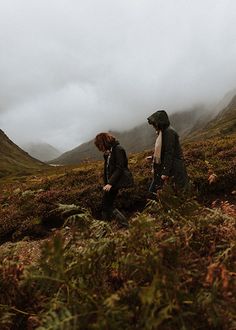 two people are walking in the grass on a cloudy day with mountains behind them and foggy skies overhead