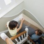 two women are painting the walls in a house with white trim and wood stairs, while another woman is sitting at the bottom of the staircase