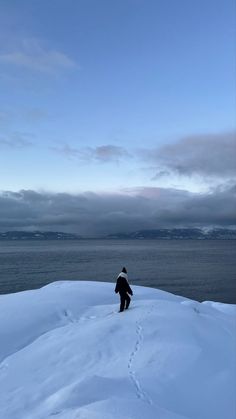 a man standing on top of a snow covered slope next to the ocean under a cloudy blue sky